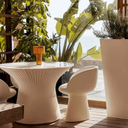 Witte, geribbelde design-eettafel met bijpassende stoel op een zonnig terras met planten en uitzicht op zee, zijaanzicht van tafel en stoel