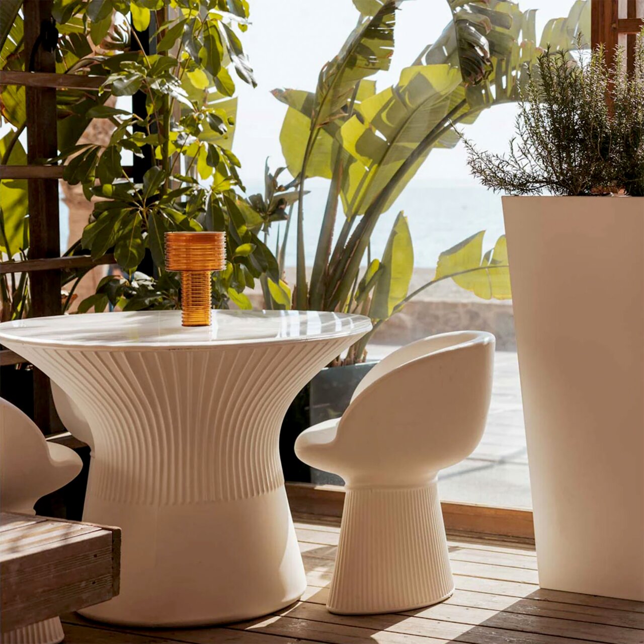 Weißer, geriffelter Design-Esstisch mit passendem Stuhl auf einer sonnigen Terrasse mit Pflanzen und Meerblick, seitliche Perspektive auf Tisch und Stuhl