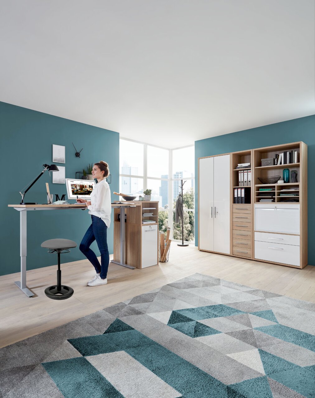 Sitness Schwinghocker / Stehhilfe CREATIV Moderner Schreibtisch mit höhenverstellbarer Funktion und Schwinghocker in einem hellen Büro, seitliche Perspektive.