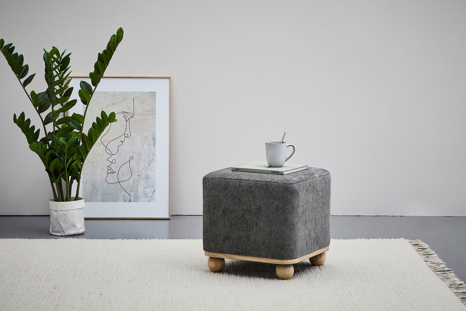 Grauer Hocker mit Holzfüßen, von der Seite fotografiert, auf einem hellen Teppich, mit einer Tasse und einem Buch darauf.