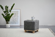 Grauer Hocker mit Holzfüßen, von der Seite fotografiert, auf einem hellen Teppich, mit einer Tasse und einem Buch darauf.