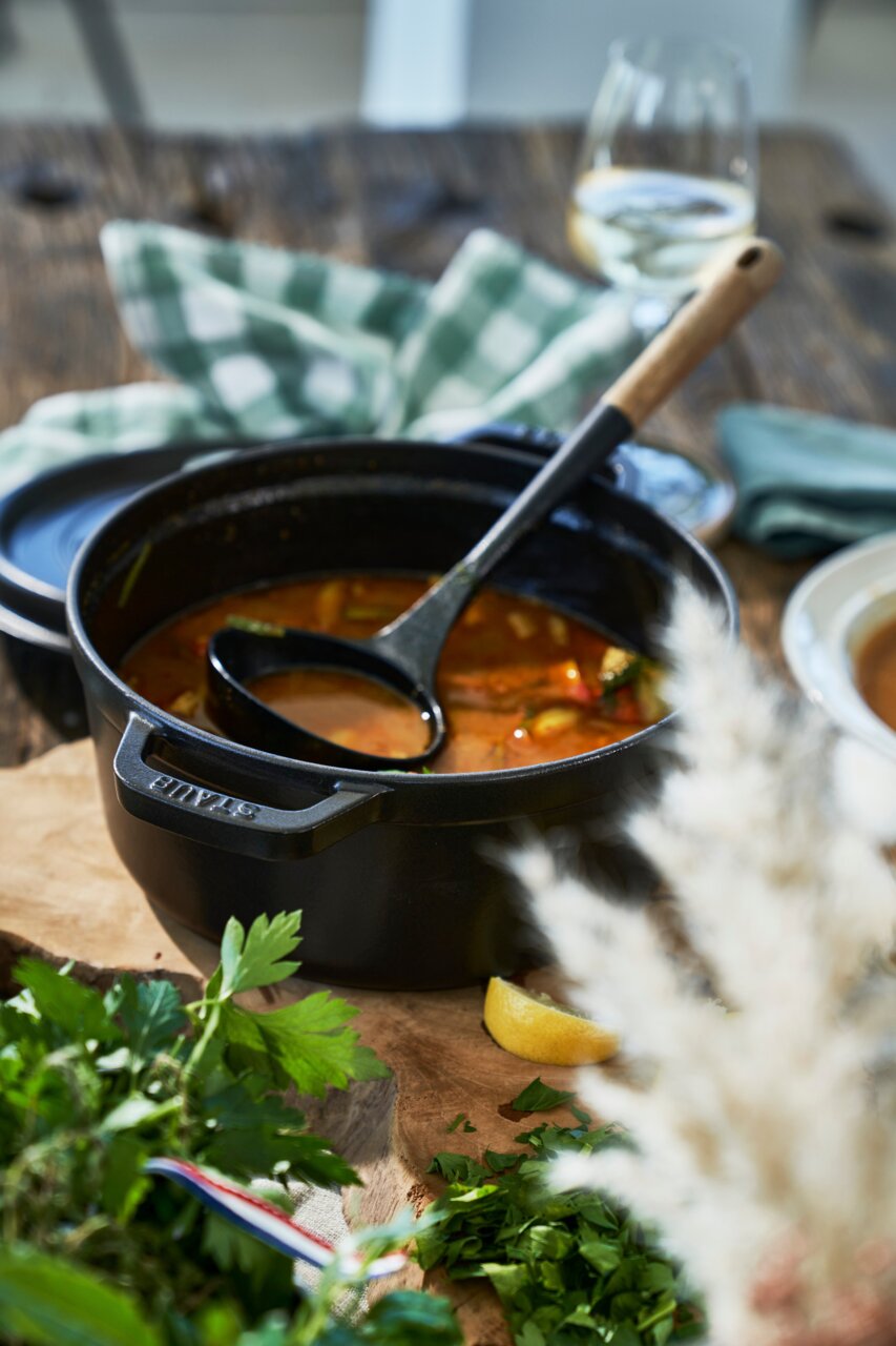 Ronde zwarte cocotte met soeplepel, gevuld met stoofpot, op een houten tafel met kruiden en citroen, gezien vanuit een zijdelingse hoek.