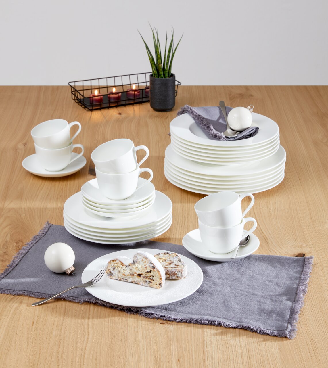 Koffieservies Noblesse, 18-delig, op een houten tafel, bestaande uit gestapelde witte kopjes en schotels, met op de achtergrond een decoratieve kaars en een plant, gefotografeerd vanuit een licht verhoogd perspectief.