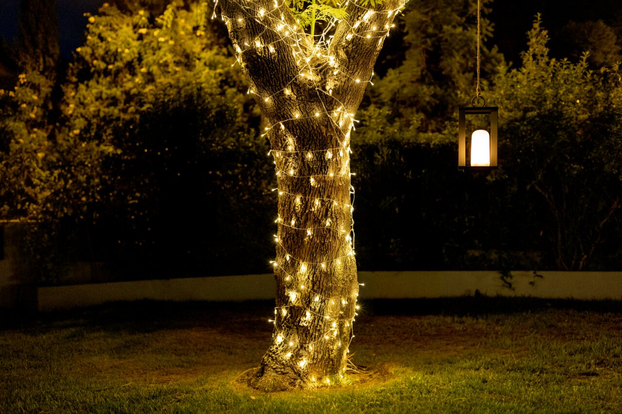 Outdoor-Lichterkette Solar, die einen Baumstamm in einem Garten bei Nacht umwickelt, mit einer hängenden Laterne im Hintergrund. Perspektive: seitlich.
