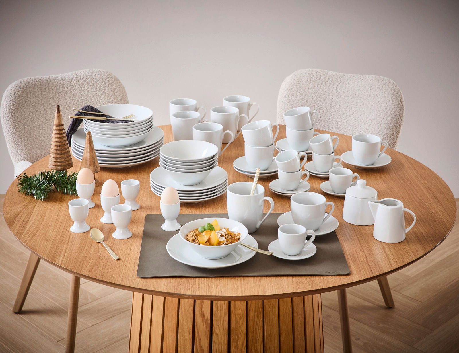 Ein 62-teiliges Kombiservice-Set auf einem runden Holztisch, bestehend aus Tellern, Tassen, Schalen und Eierbechern in schlichtem Weiß. Die Perspektive zeigt das Set von oben, mit zwei Stühlen im Hintergrund.