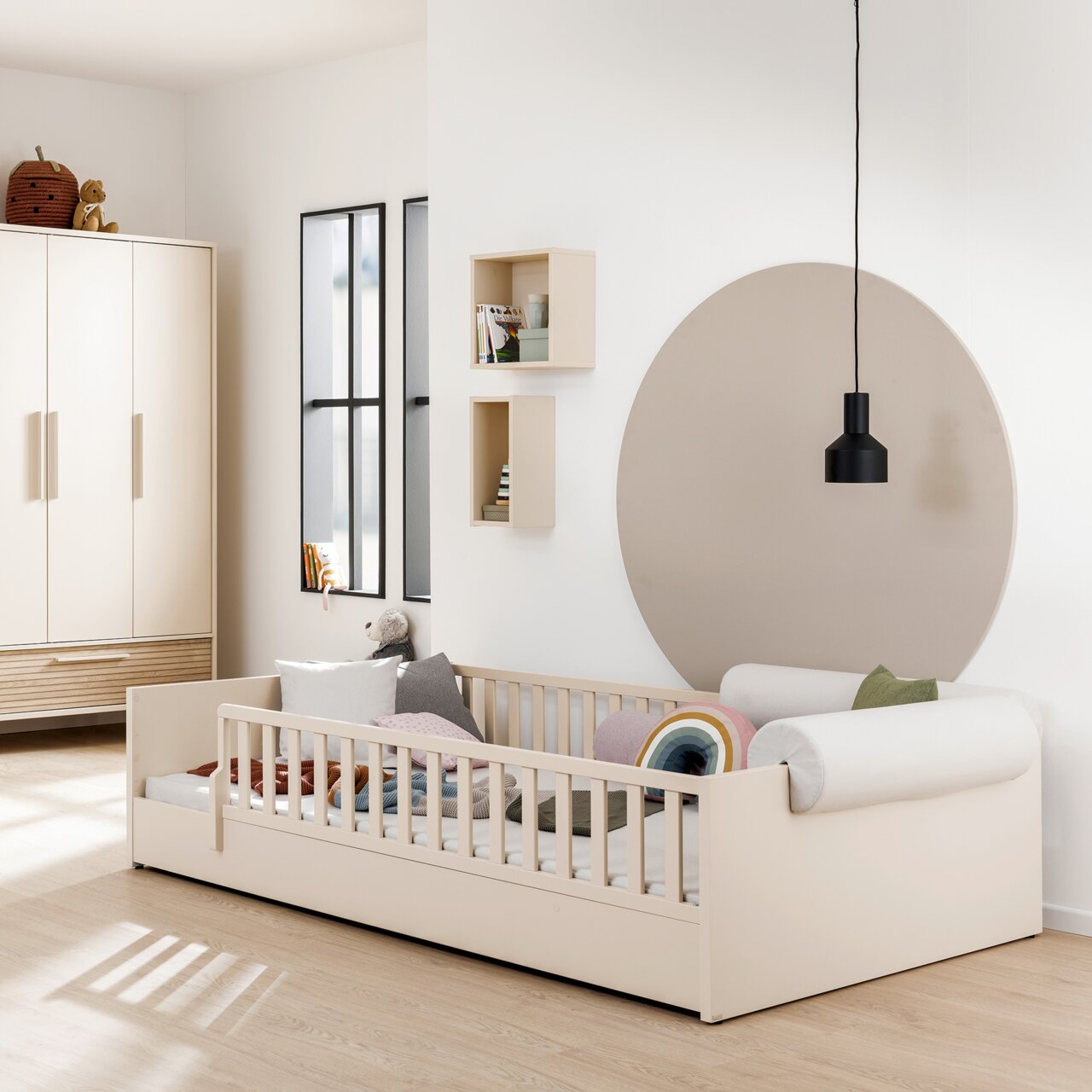 Paidi Bodenbett LITTLE FLO Kinderzimmer mit einem cremefarbenen Bodenbett mit Rausfallschutz, dekoriert mit bunten Kissen und Decken. Im Hintergrund sind ein Kleiderschrank, Regale und eine runde Wanddekoration zu sehen. Perspektive von der Seite.
