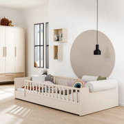 Kinderzimmer mit einem cremefarbenen Bodenbett mit Rausfallschutz, dekoriert mit bunten Kissen und Decken. Im Hintergrund sind ein Kleiderschrank, Regale und eine runde Wanddekoration zu sehen. Perspektive von der Seite.