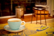 Hellblaue Keramik-Kaffeetasse mit Untertasse auf gelbem Tischläufer, perspektivisch aus leicht erhöhter Frontansicht mit unscharfem Café-Hintergrund.