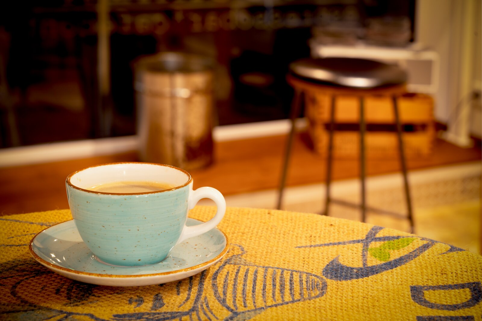 Hellblaue Keramik-Kaffeetasse mit Untertasse auf gelbem Tischläufer, perspektivisch aus leicht erhöhter Frontansicht mit unscharfem Café-Hintergrund.