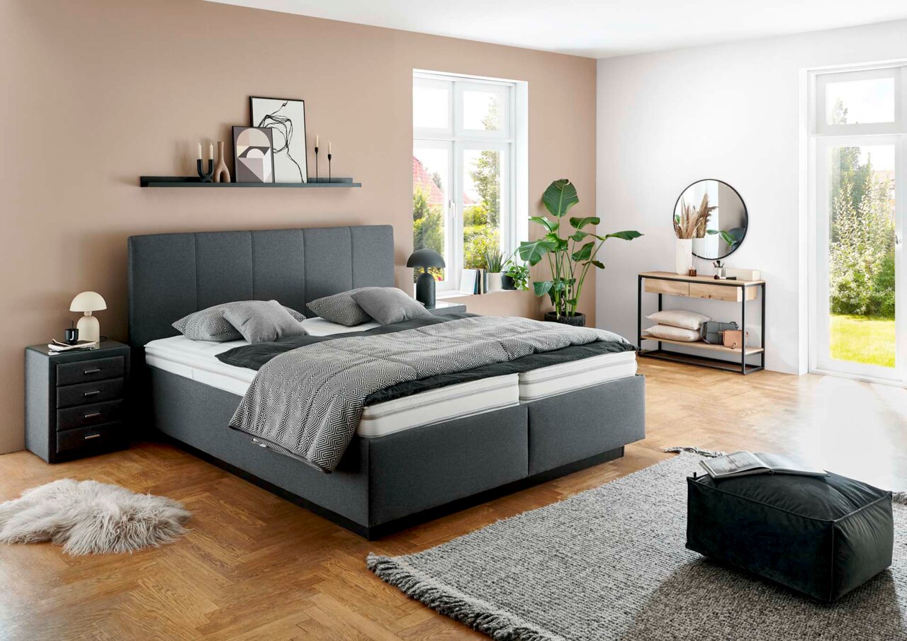 Modernes graues Polsterbett in einem stilvoll eingerichteten Schlafzimmer mit Holzboden, Pflanzen und großen Fenstern, seitliche Perspektive.