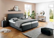 Modernes graues Polsterbett in einem stilvoll eingerichteten Schlafzimmer mit Holzboden, Pflanzen und großen Fenstern, seitliche Perspektive.