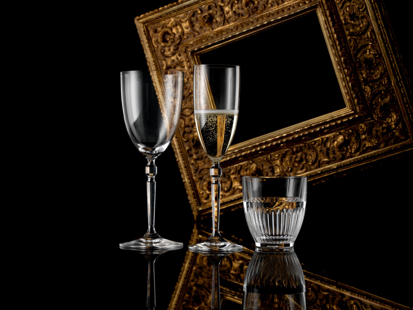 Drei elegante Gläser (zwei hohe Sekt-/Weingläser, eines mit prickelndem Getränk, und ein geriffeltes Tumblerglas) vor einem goldverzierten Bilderrahmen auf schwarzem, spiegelndem Untergrund, fotografiert in leichter Frontansicht