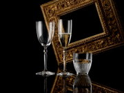Drei elegante Gläser (zwei hohe Sekt-/Weingläser, eines mit prickelndem Getränk, und ein geriffeltes Tumblerglas) vor einem goldverzierten Bilderrahmen auf schwarzem, spiegelndem Untergrund, fotografiert in leichter Frontansicht