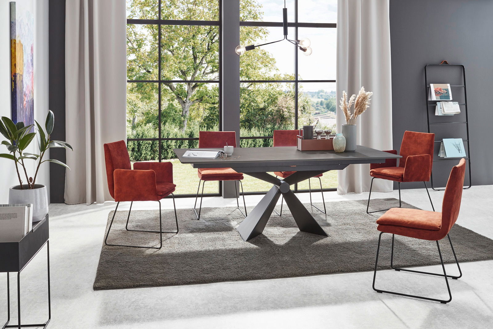 Moderner Esstisch mit schwarzer Tischplatte und X-förmigem Gestell, umgeben von roten Stühlen in einem hellen Raum mit großen Fenstern, seitliche Perspektive.
