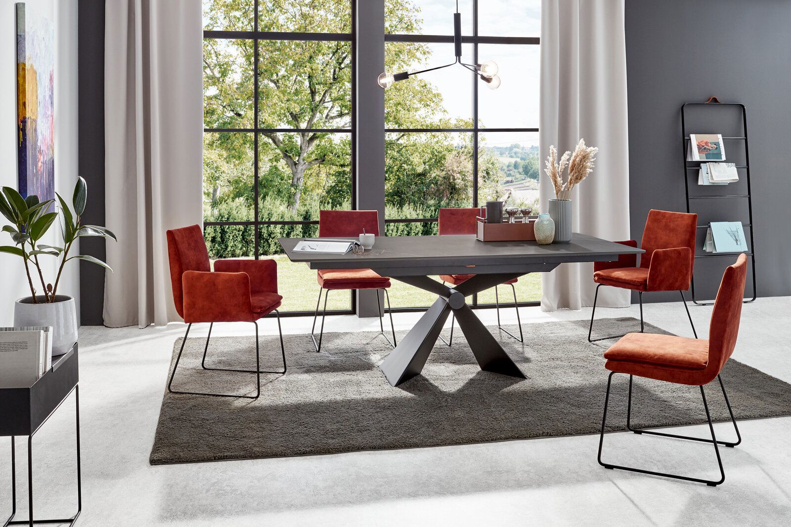 Moderner Esstisch mit schwarzer Tischplatte und X-förmigem Gestell, umgeben von roten Stühlen in einem hellen Raum mit großen Fenstern, seitliche Perspektive.