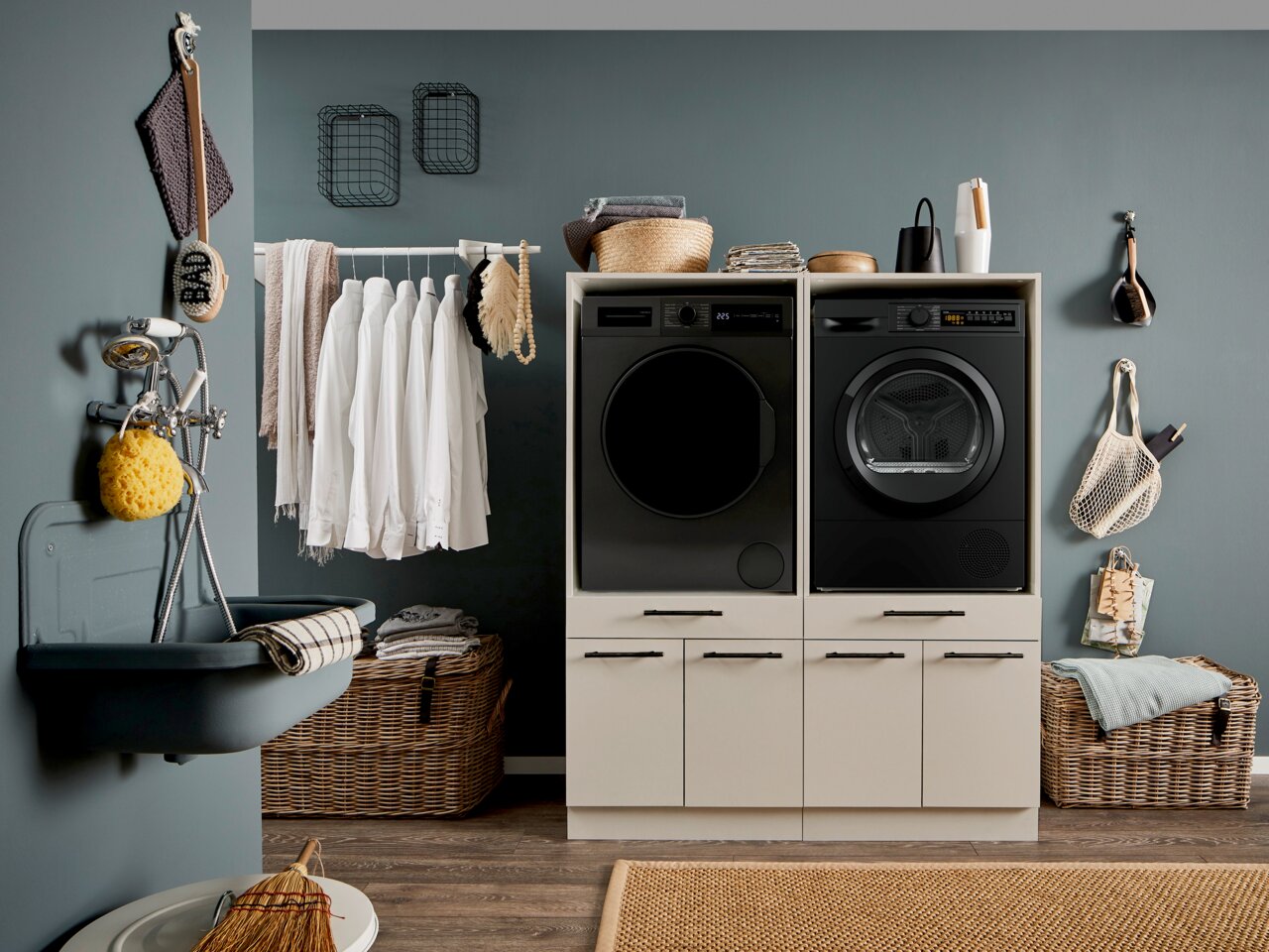 Modern eingerichteter Hauswirtschaftsraum mit schwarzer Waschmaschine und schwarzem Trockner in weißem Unterschrank, Wäschekorb und aufgehängten Hemden; frontale Perspektive auf die Geräte.