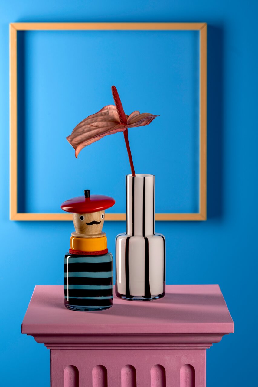 Vorderansicht einer stilvollen Objektvase mit Pizzaiolo-Motiv, neben einer modernen, metallisch glänzenden Vase mit einer exotischen Blume, auf einem pinkfarbenen Sockel vor einem blauen Hintergrund.