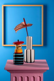 Vorderansicht einer stilvollen Objektvase mit Pizzaiolo-Motiv, neben einer modernen, metallisch glänzenden Vase mit einer exotischen Blume, auf einem pinkfarbenen Sockel vor einem blauen Hintergrund.