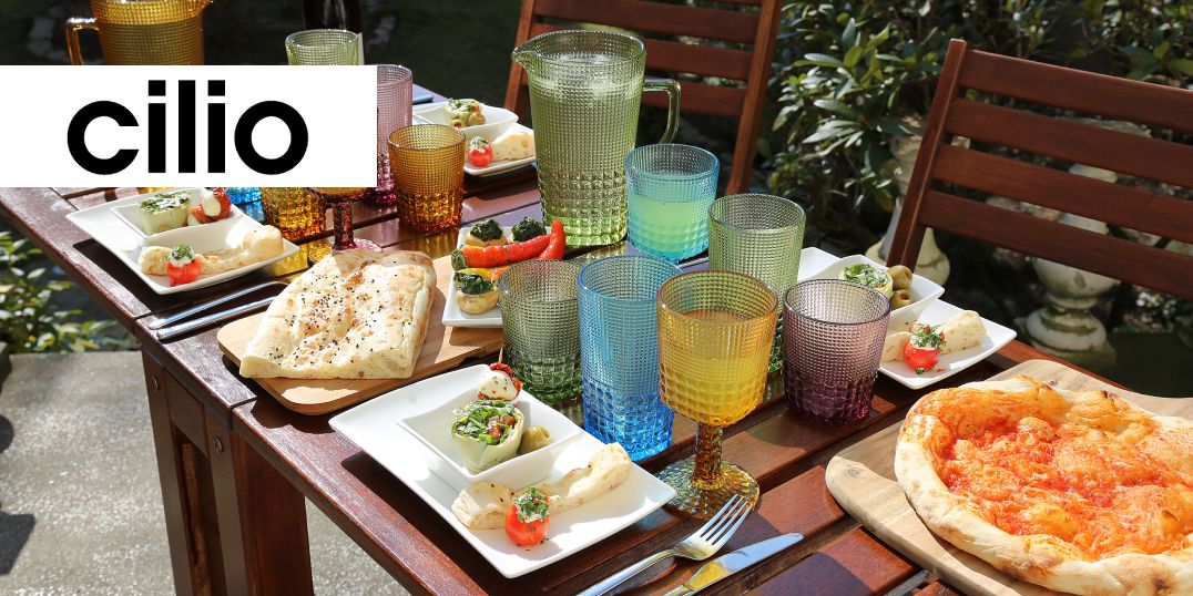 Ein buntes Esszimmer im Garten, mit bunten Gläsern, Pizzen und Vorspeisen auf einem Holztisch. Logo Cilio