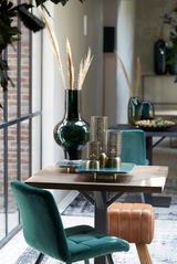 Moderner Innenraum mit einem grünen Samtstuhl und einem braunen Lederhocker an einem Holztisch, dekoriert mit einer großen grünen Vase und Kerzenhaltern. Perspektive von der Seite.