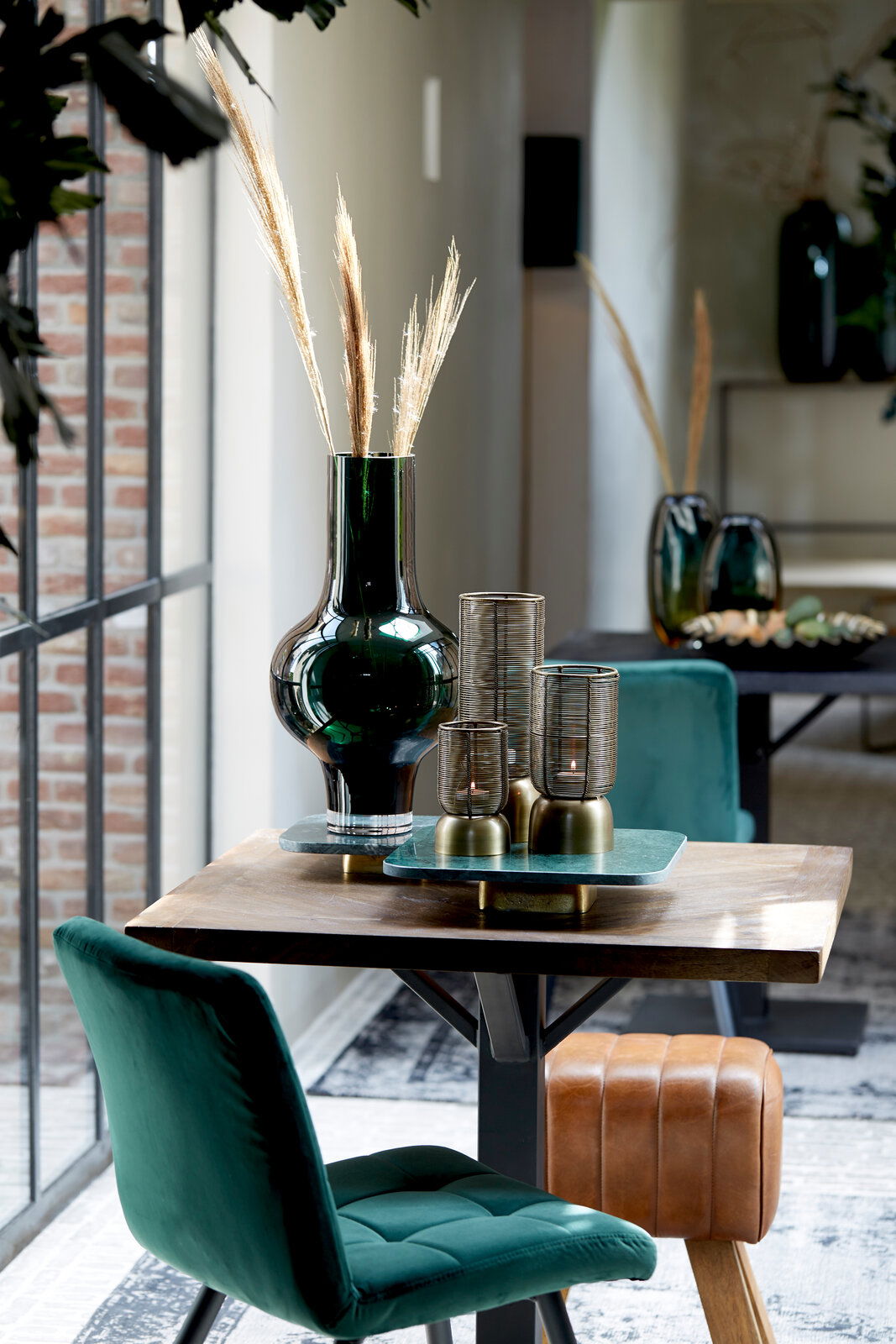 Moderner Innenraum mit einem grünen Samtstuhl und einem braunen Lederhocker an einem Holztisch, dekoriert mit einer großen grünen Vase und Kerzenhaltern. Perspektive von der Seite.