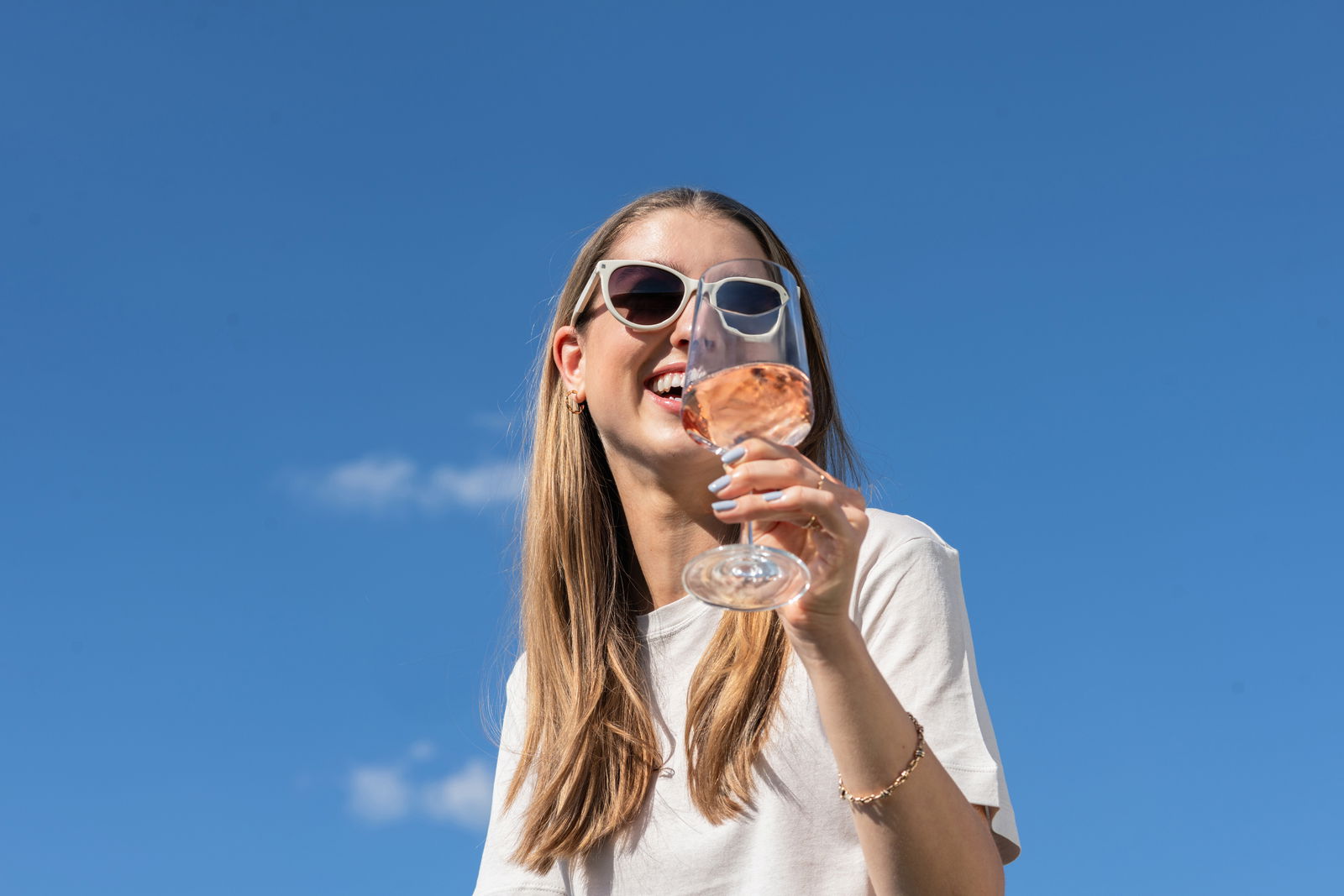 Frau mit Sonnenbrille hält ein Weißweinglas in der Hand, blauer Himmel im Hintergrund, Perspektive von unten.