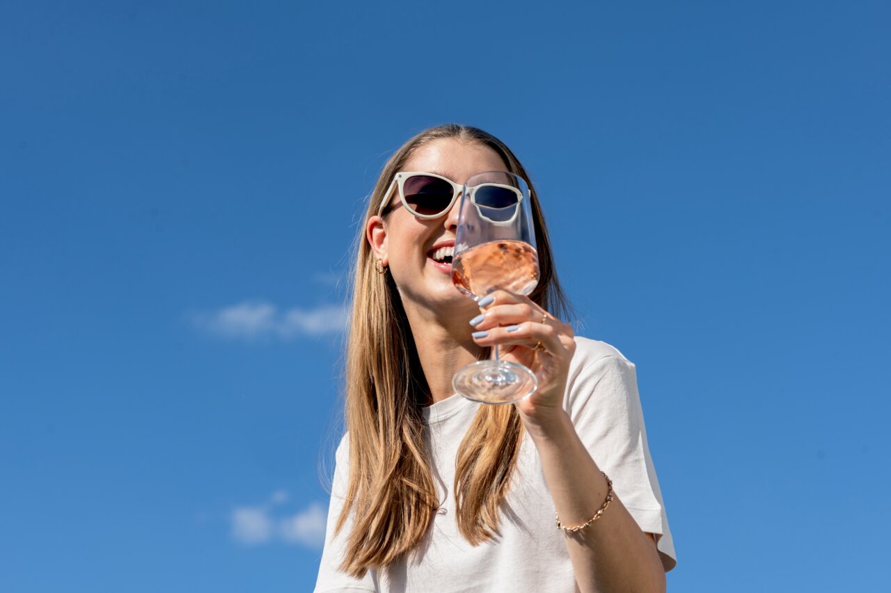 Frau mit Sonnenbrille hält ein Weißweinglas in der Hand, blauer Himmel im Hintergrund, Perspektive von unten.
