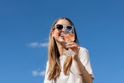 Een vrouw met een zonnebril houdt een glas witte wijn vast, met een blauwe lucht op de achtergrond, gefotografeerd vanuit een laag standpunt.