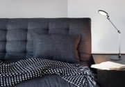 Moderne Tischleuchte auf einem Nachttisch neben einem Sofa mit grauem Kissen und schwarz-weiß gemustertem Überwurf, seitliche Perspektive.