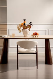 Frontale Ansicht: Moderner Esszimmerstuhl mit cremefarbenem Bouclé-Bezug und dunklem Holzgestell vor einem Holztisch in hellem, minimalistischen Interieur.