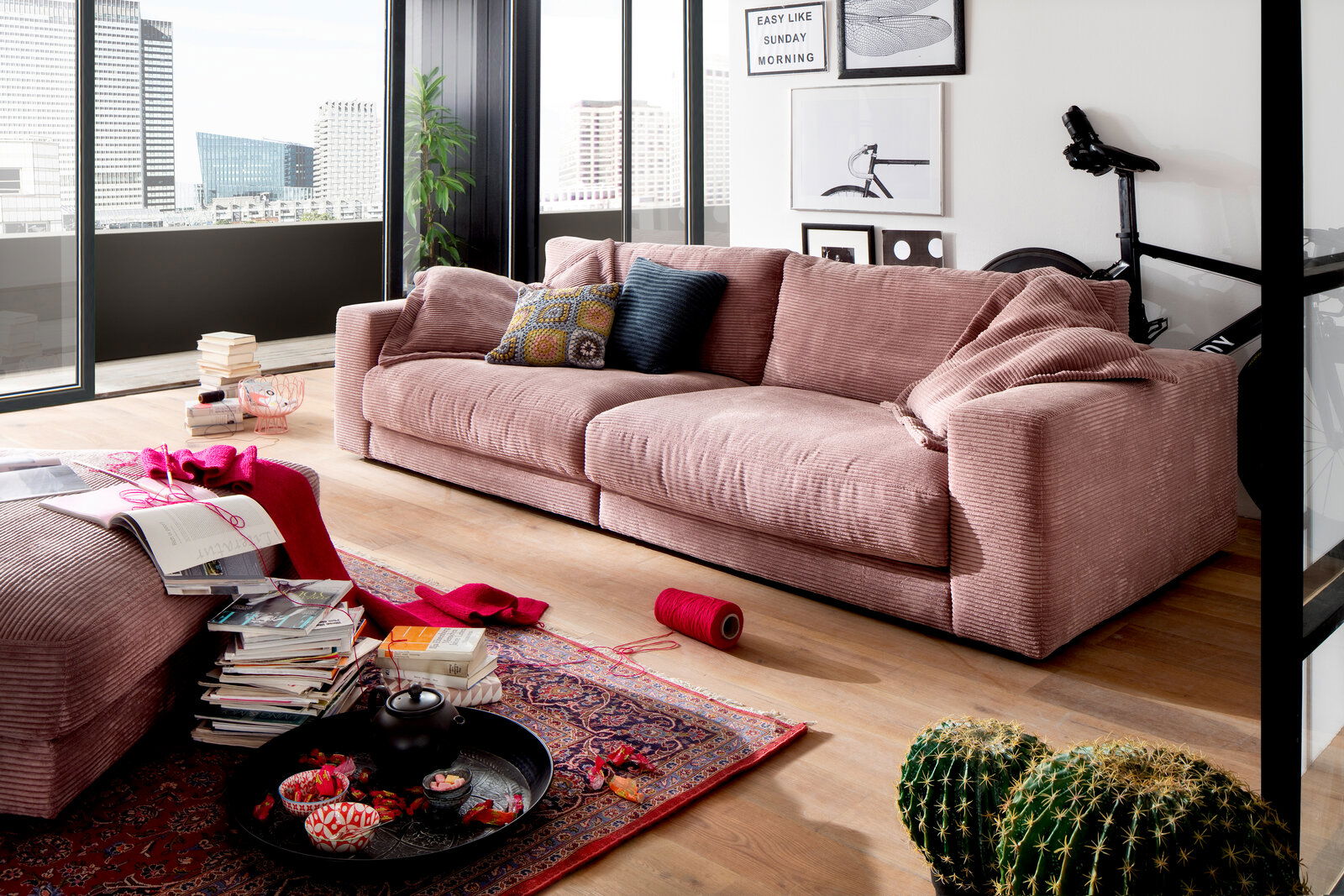 Gemütliches rosa Sofa in einem modernen Wohnzimmer mit großen Fenstern, dekoriert mit Kissen und Decken, seitliche Perspektive.