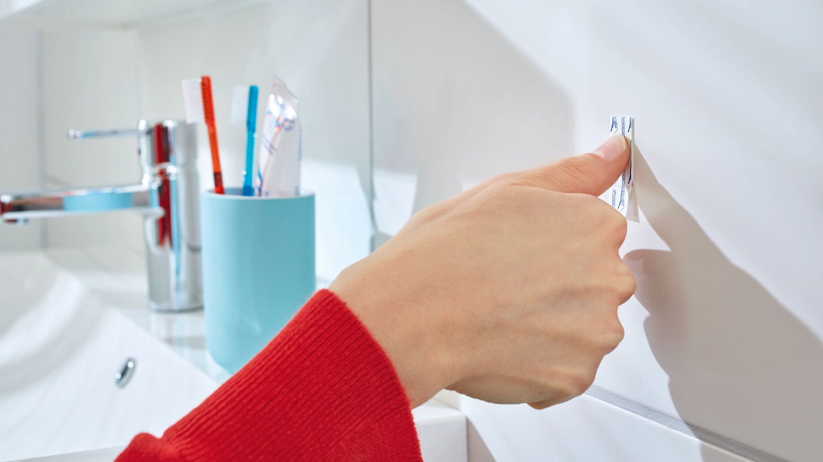 Nahaufnahme einer Hand, die einen tesa Powerstrip an einer weißen Badezimmerwand anbringt. Im Hintergrund sind ein Wasserhahn und ein Zahnputzbecher zu sehen.