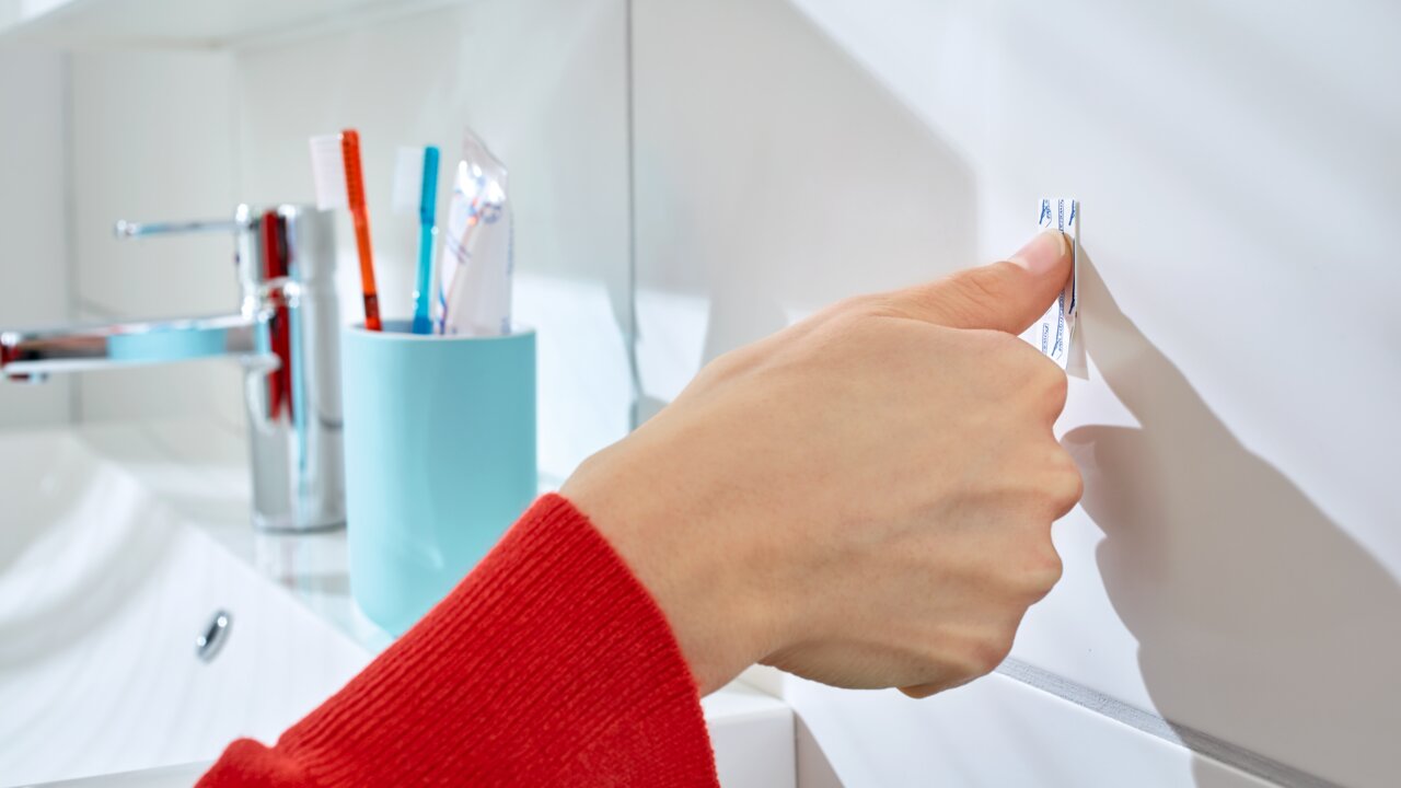 Nahaufnahme einer Hand, die einen tesa Powerstrip an einer weißen Badezimmerwand anbringt. Im Hintergrund sind ein Wasserhahn und ein Zahnputzbecher zu sehen.