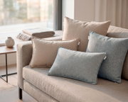 Beige- und blau-graue Dekokissen auf einem hellgrauen Sofa am Fenster, aufgenommen in schräger Vorderansicht (3/4-Perspektive)