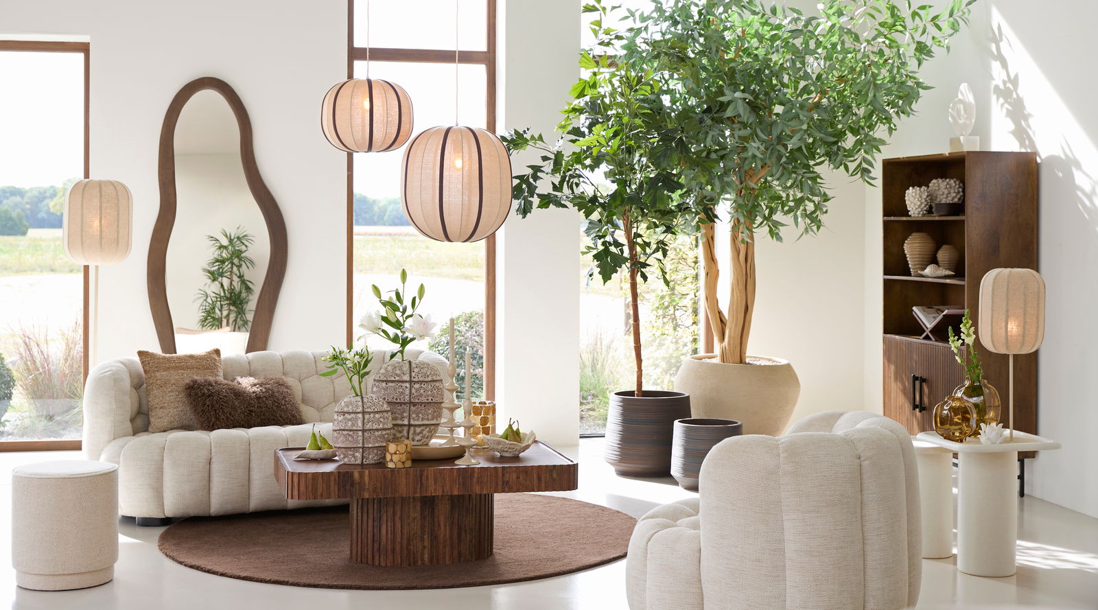 Gemütliches Wohnzimmer mit beigem Sofa und Kissen, umgeben von Pflanzen und stilvollen Lampen, aus der Perspektive eines offenen Raumes mit großen Fenstern.