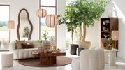Gemütliches Wohnzimmer mit beigem Sofa und Kissen, umgeben von Pflanzen und stilvollen Lampen, aus der Perspektive eines offenen Raumes mit großen Fenstern.