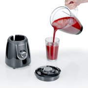 Severin blenderbasis met draaiknop en losse deksel op witte achtergrond; kan met rood smoothie-achtig drankje wordt vanuit een schenkkan in een glas gegoten, schuin van voren gefotografeerd