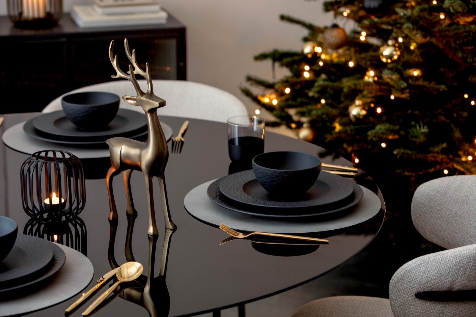 Goldfarbene Hirschfigur aus leicht seitlicher Vorderansicht als festliche Tischdekoration auf einer schwarz gedeckten Tafel vor einem beleuchteten Weihnachtsbaum