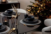 Goldfarbene Hirschfigur aus leicht seitlicher Vorderansicht als festliche Tischdekoration auf einer schwarz gedeckten Tafel vor einem beleuchteten Weihnachtsbaum