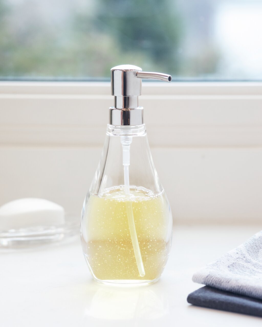 Transparenter Seifenspender mit silbernem Pumpkopf, gefüllt mit gelber Flüssigseife, auf einer Fensterbank von vorne fotografiert.