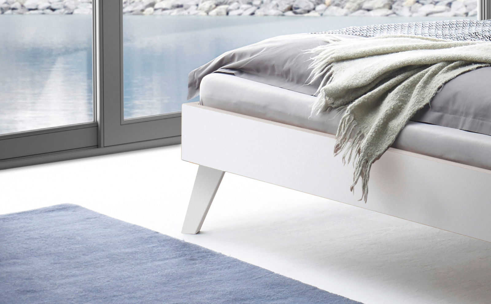 Modernes weißes Bettgestell mit schrägen Beinen, seitliche Perspektive, mit grauer Decke und Kissen, vor einem großen Fenster mit Seeblick.