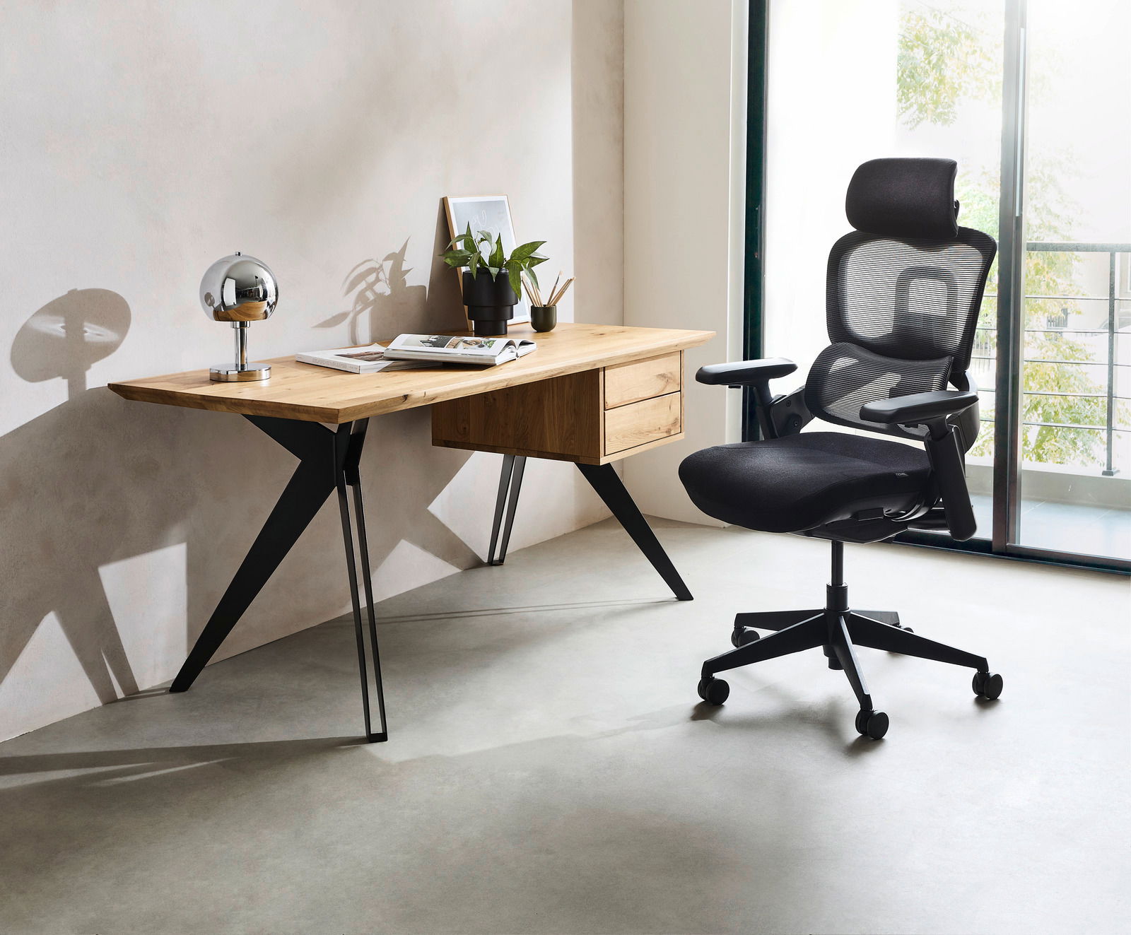 Moderner Schreibtisch aus Holz mit schwarzen Metallbeinen, seitlich fotografiert, neben einem ergonomischen Bürostuhl mit Netzrückenlehne.