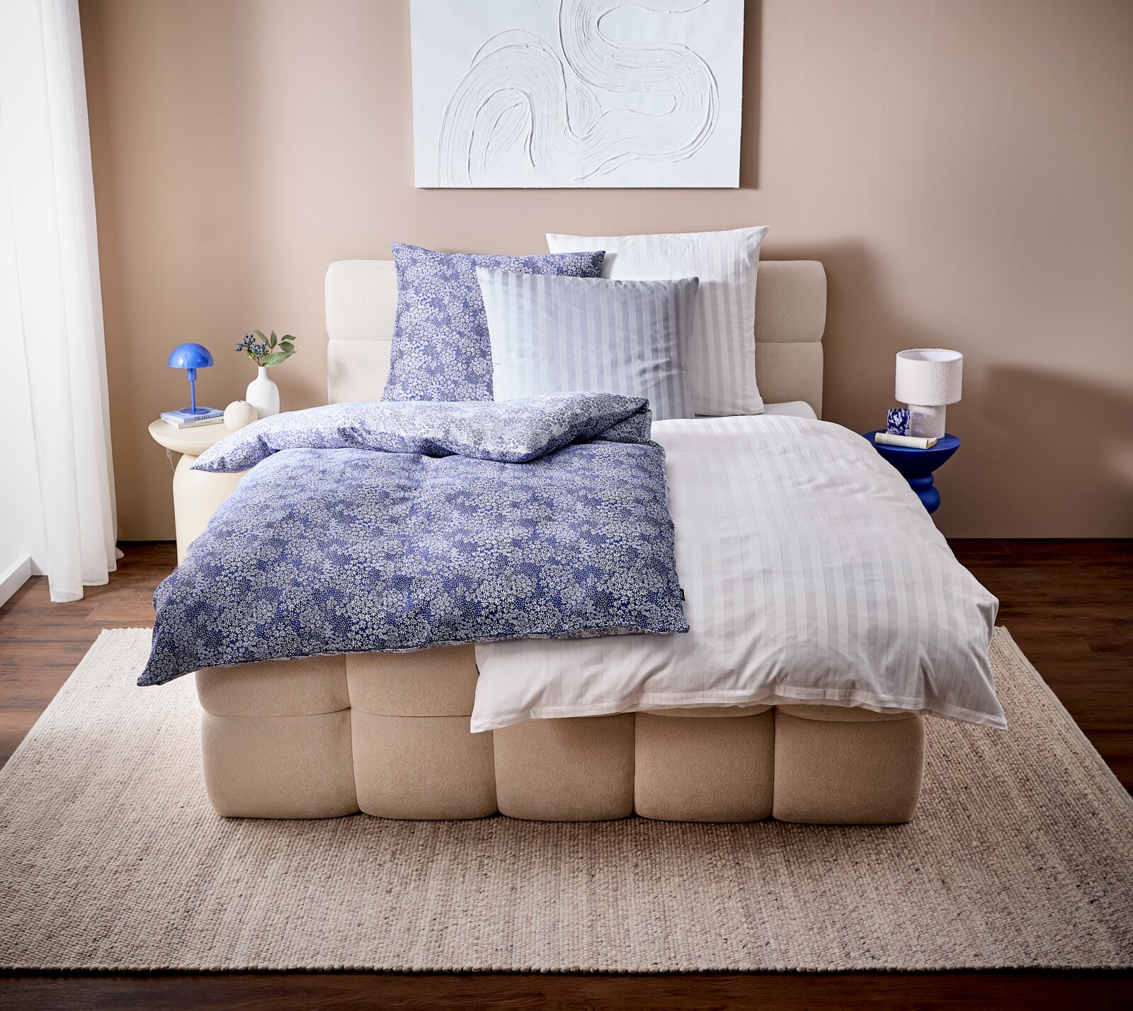 Modernes Schlafzimmer mit beigem Polsterbett und Bettwäsche-Set: blaue, fein gemusterte Decke und Kissen kombiniert mit hellgrau-weiß gestreifter Bettwäsche; Perspektive: frontale Ansicht leicht von oben.