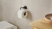 Schwarzer Toilettenpapierhalter der Serie tesa MOON BLACK, montiert an einer Badezimmerwand mit einer Rolle Toilettenpapier, seitliche Perspektive.