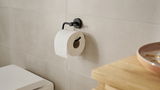Schwarzer Toilettenpapierhalter der Serie tesa MOON BLACK, montiert an einer Badezimmerwand mit einer Rolle Toilettenpapier, seitliche Perspektive.