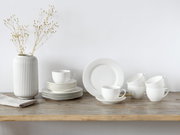 Minimalistisches weißes Geschirr-Set mit Tellern und Tassen auf rustikalem Holztisch neben geriffelter weißer Vase mit Zweigen, frontal in leichter Seitenansicht fotografiert.