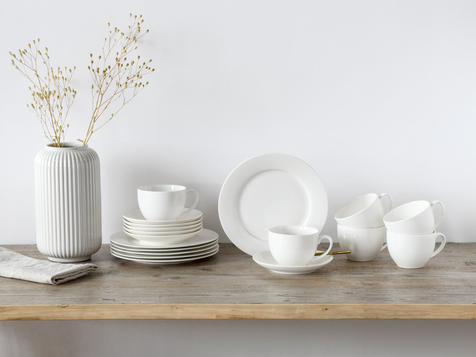 Minimalistisches weißes Geschirr-Set mit Tellern und Tassen auf rustikalem Holztisch neben geriffelter weißer Vase mit Zweigen, frontal in leichter Seitenansicht fotografiert.