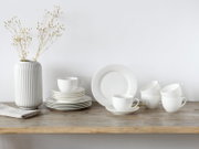 Minimalistisches weißes Geschirr-Set mit Tellern und Tassen auf rustikalem Holztisch neben geriffelter weißer Vase mit Zweigen, frontal in leichter Seitenansicht fotografiert.