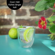 Doppelwandiges Glas mit Limettenscheiben und Minze, auf einem türkisfarbenen Tisch, mit Ziegelwand und Pflanzen im Hintergrund. Perspektive: seitlich.