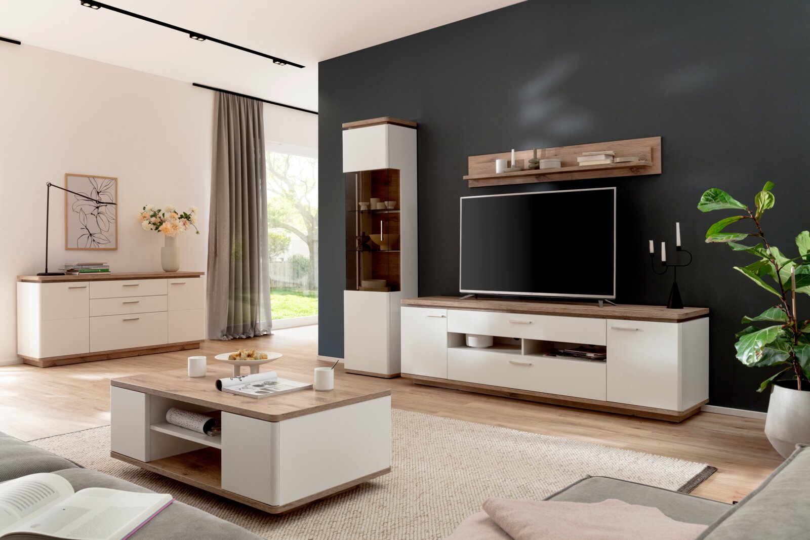 Modernes Wohnzimmer mit weißer Wohnwand und Couchtisch in Holzoptik, TV-Board mit Fernseher, Vitrine und Sideboard vor dunkler Akzentwand, aufgenommen aus schräger Frontperspektive auf Augenhöhe.